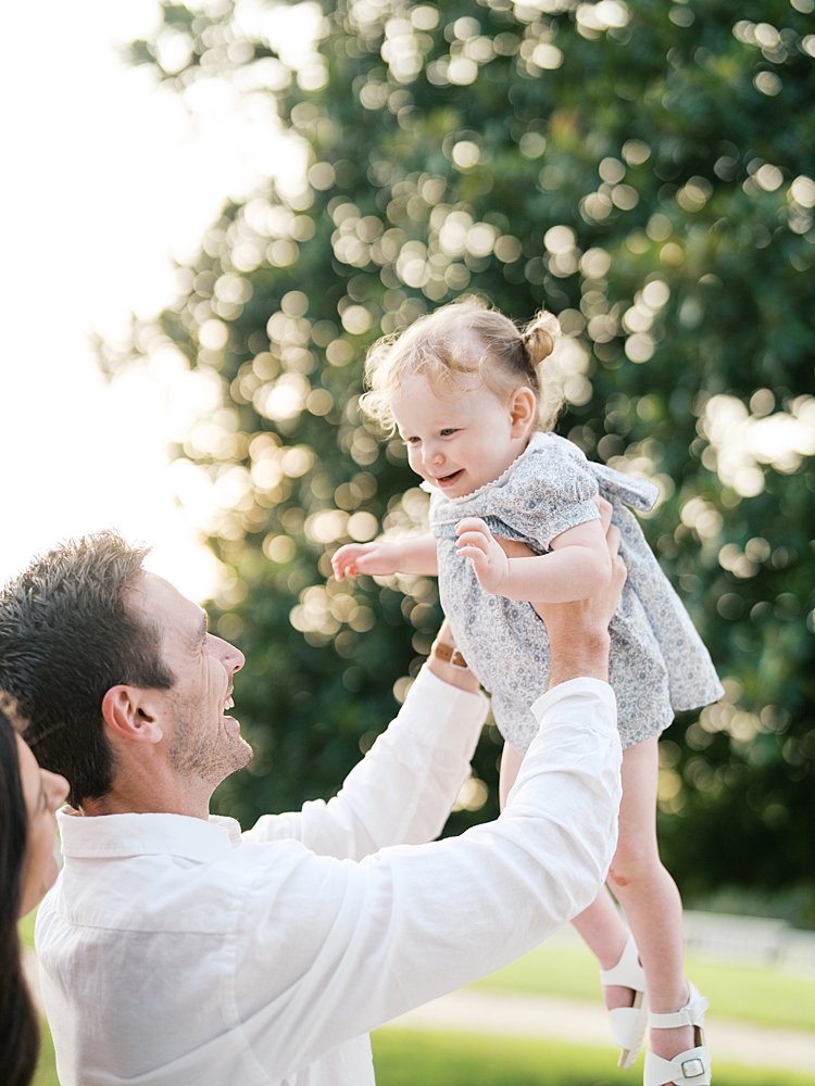 A father holds his toddler daughter up in the air as she smiles.