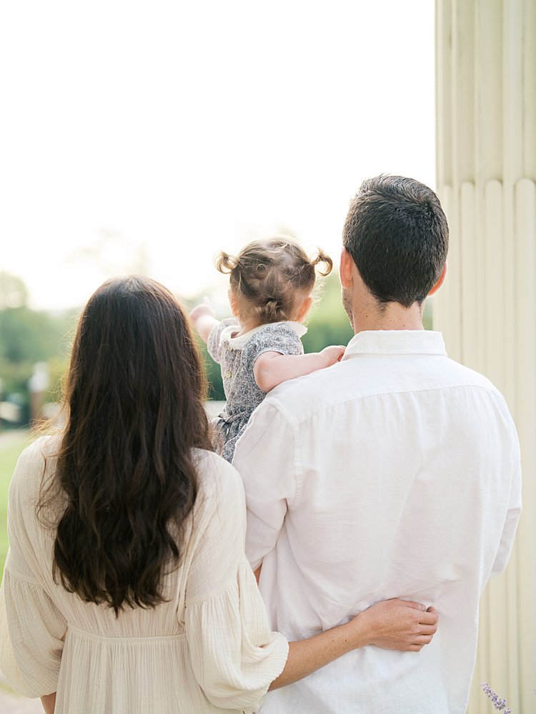 Mother, father, and their toddler daughter stand on the porch of Oatlands historic home and look out onto the grounds.