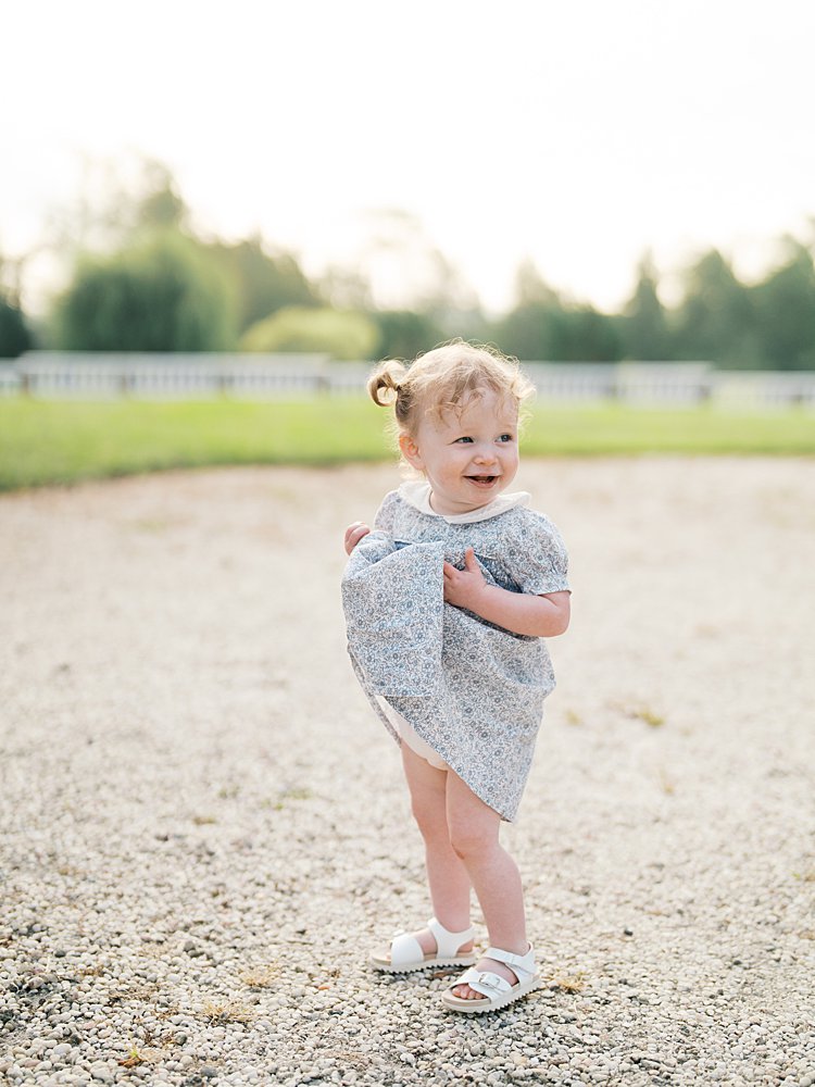 A toddler girl with pigtails holds up her dress and smiles.