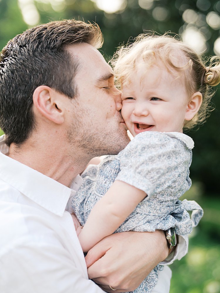 A father leans in to kiss his toddler daughter's cheek.