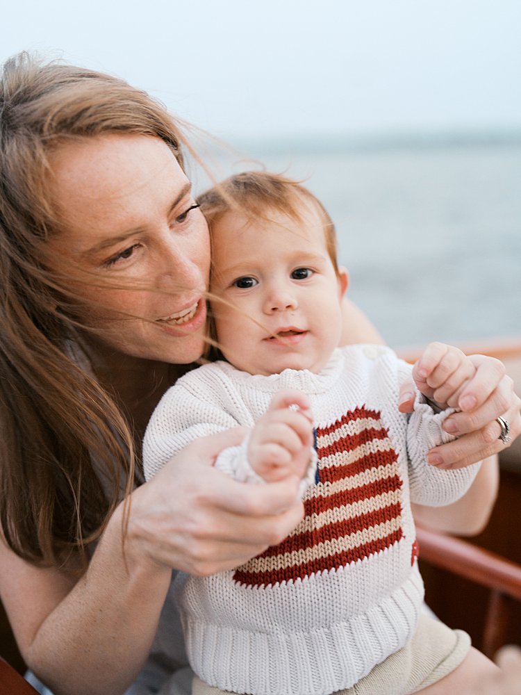 Red-haired mother holds her baby boy's hands as he looks at the camera with an American flag on his sweater.