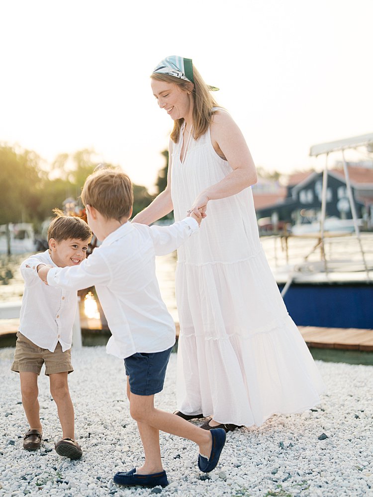 Mother in white dress and green bandana dances with her two young sons at a dock.