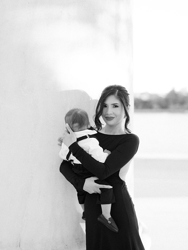 A Mother In A Black Dress Holds Her Baby Boy While Leaning Against A Column At The Jefferson Memorial.
