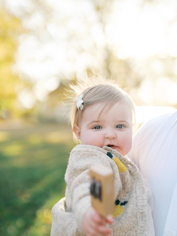 A Baby Girl Soaked In Sunlight Holds A Rattle Up To The Camera.