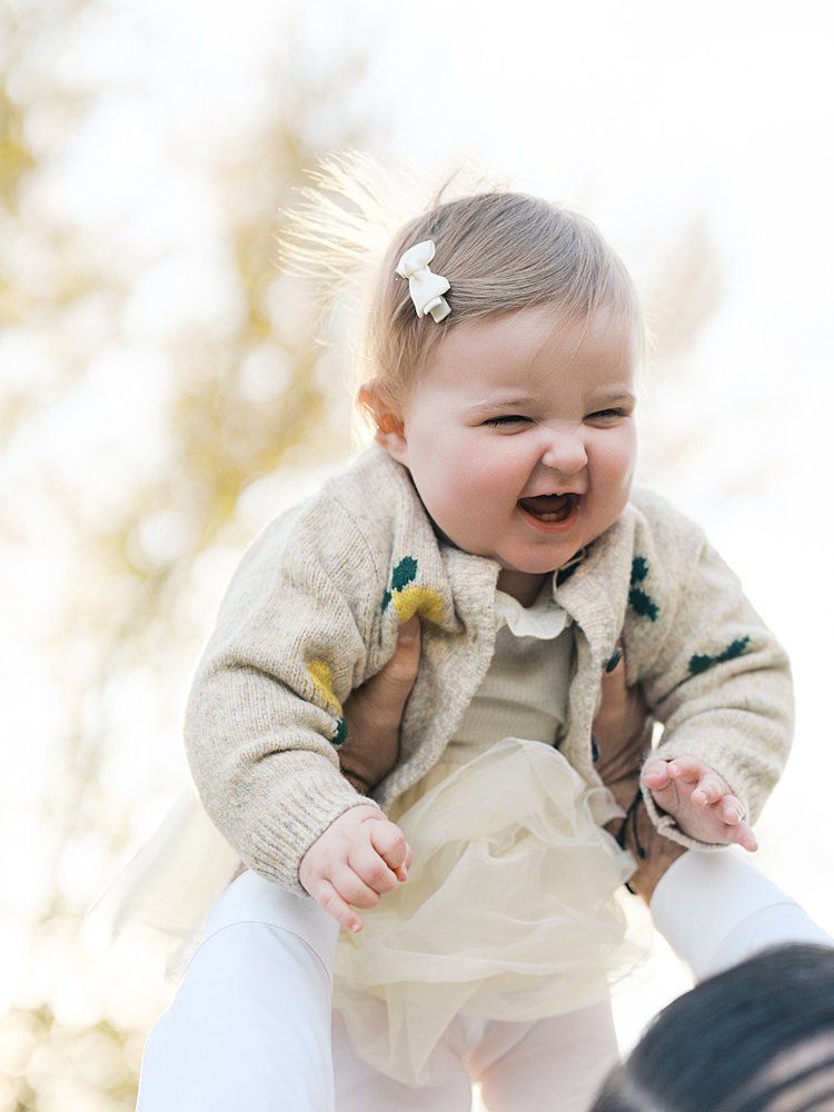 A Baby Girl Is Held Up In The Air And Gives A Big Smile.
