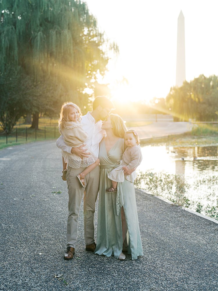 A Father Kisses His Wife's Head As They Hold Their Two Young Girls During Their Downtown Dc Family Photos At Constitution Gardens.