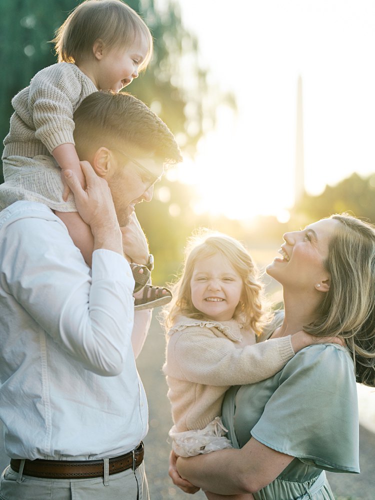 Mother And Father Smile And Laugh As They Hold Their Two Young Girls During Their Downtown Dc Family Photos At Constitution Gardens.