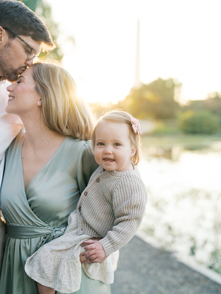 Little Girl Smiles At The Camera While Her Dad Kisses Her Mom's Head.