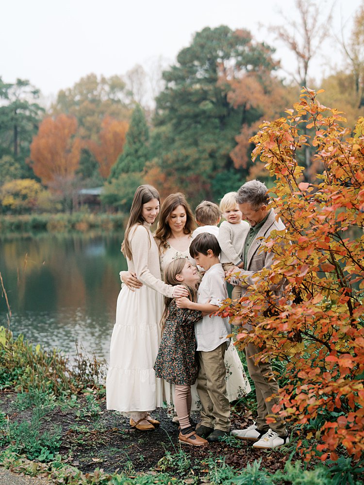 A Family Of Seven Stands Along A Lake Near An Orange-Leave Tree.
