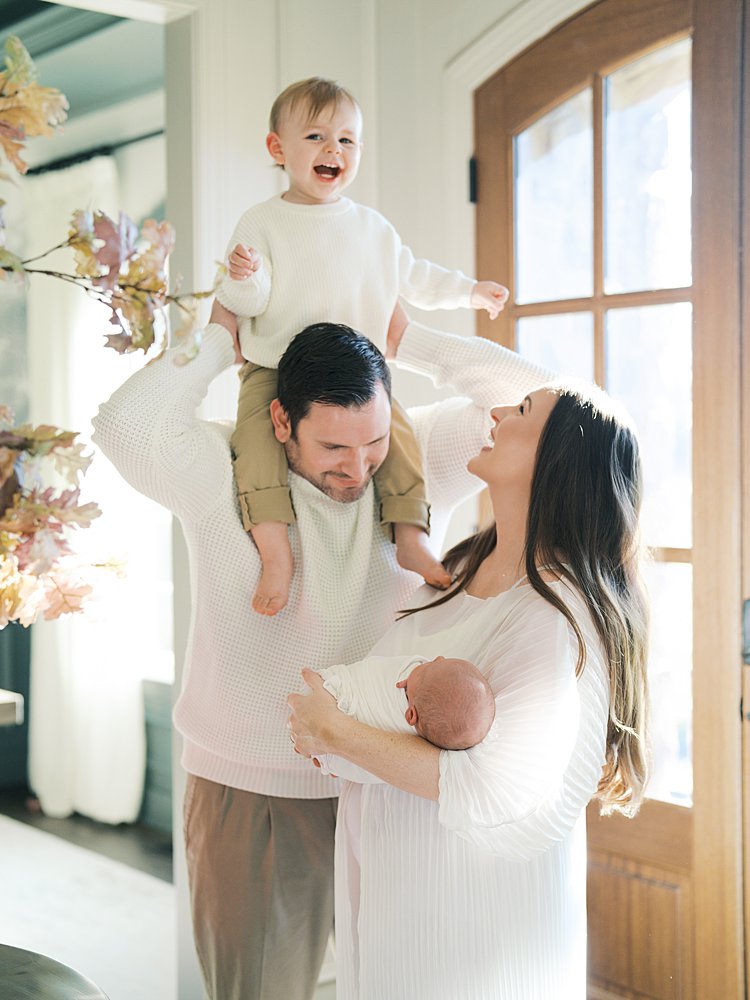 Toddler On Dad's Shoulders Laughs As Mother Looks Up At Him While Holding Newborn Baby.