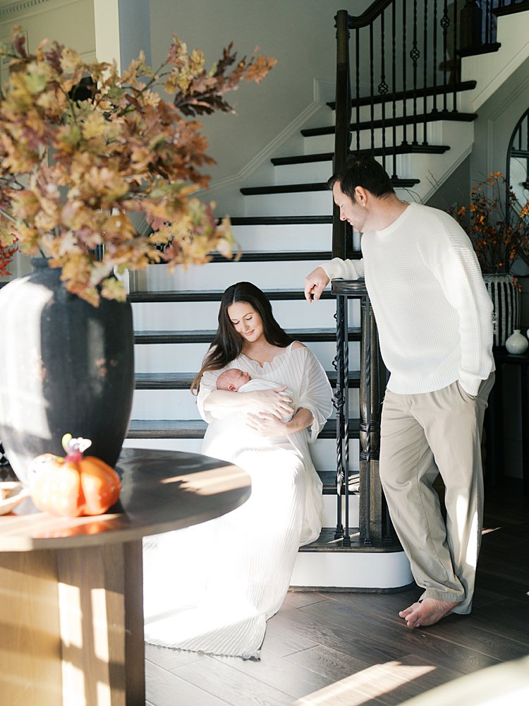 Father Leans On Staircase Looking Down At His Wife, Sitting On The Stairs And Holding Their Newborn During Their Brookeville Md Newborn Photos.