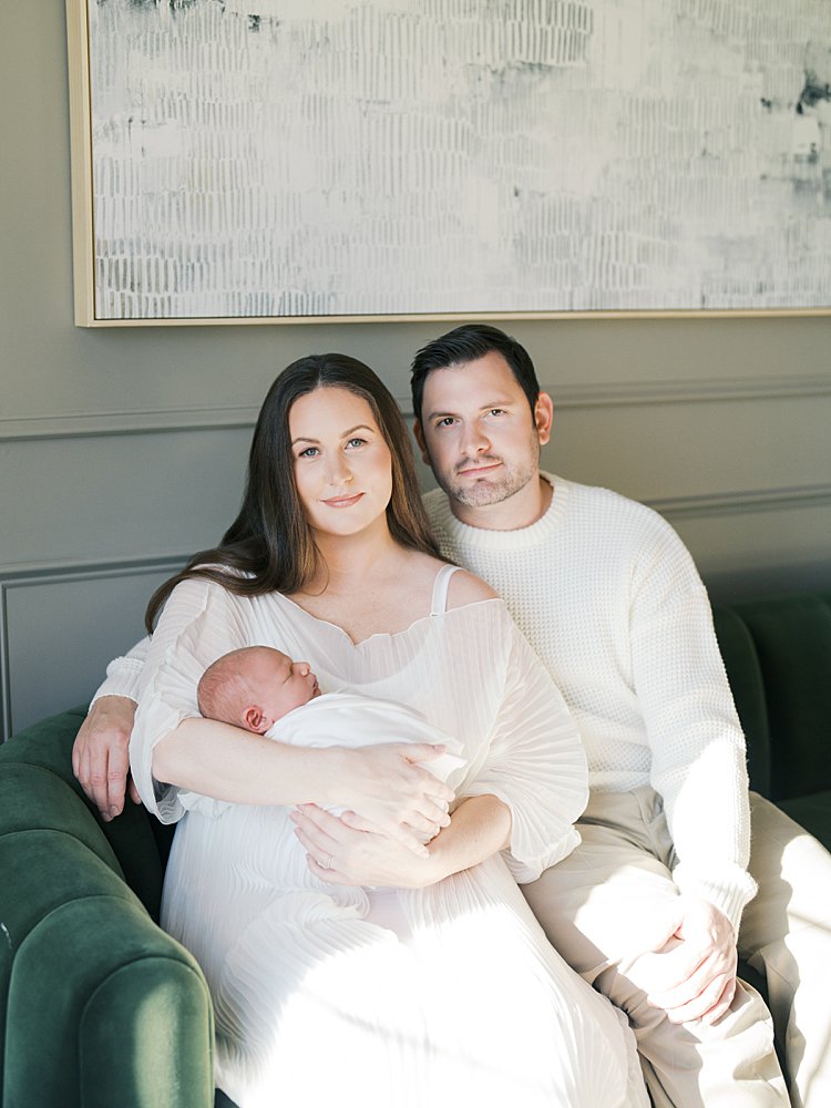 Mother And Father Look Directly At The Camera With Soft Smiles While Sitting On A Green Velvet Couch During Their Brookeville Md Newborn Photos.