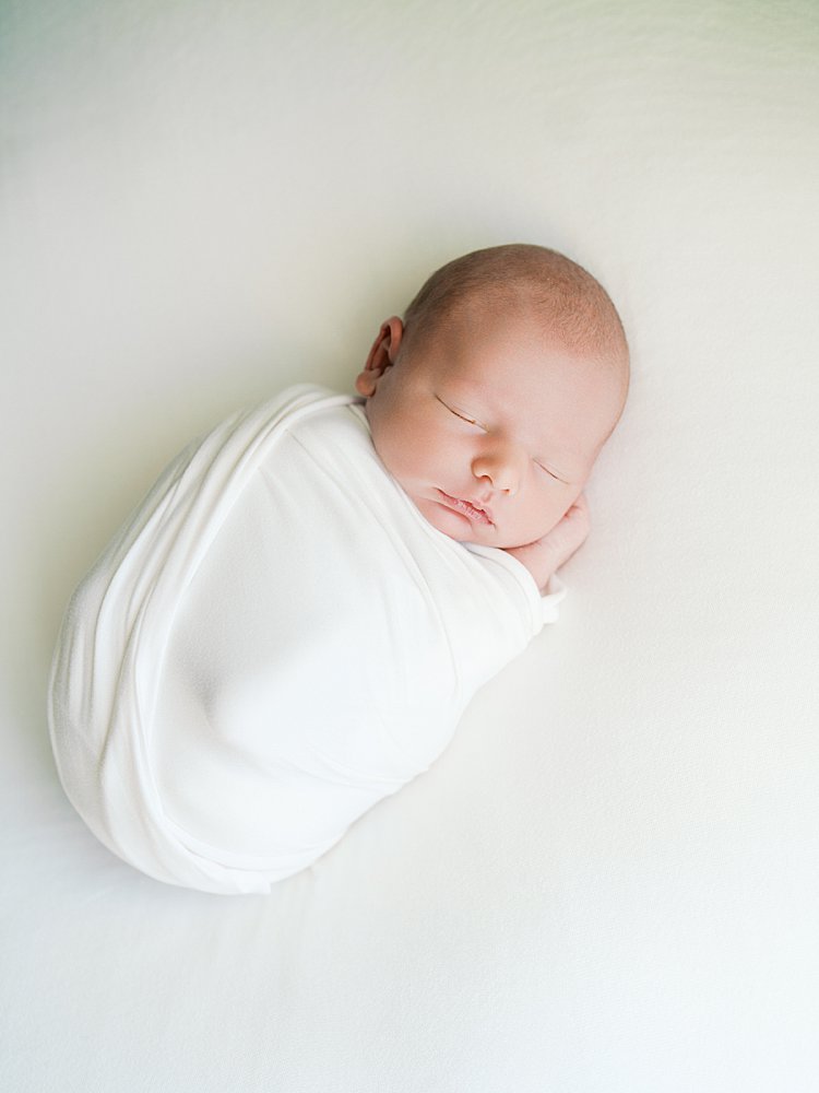 Newborn Baby Wrapped In White Swaddles Sleeps Peacefully On A Bed.