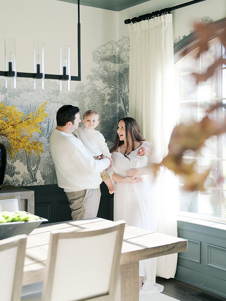Mother Smiles At Her Toddler Son Held By Her Husband As They Stand In Their Dining Room During Their Brookeville Md Newborn Photos.