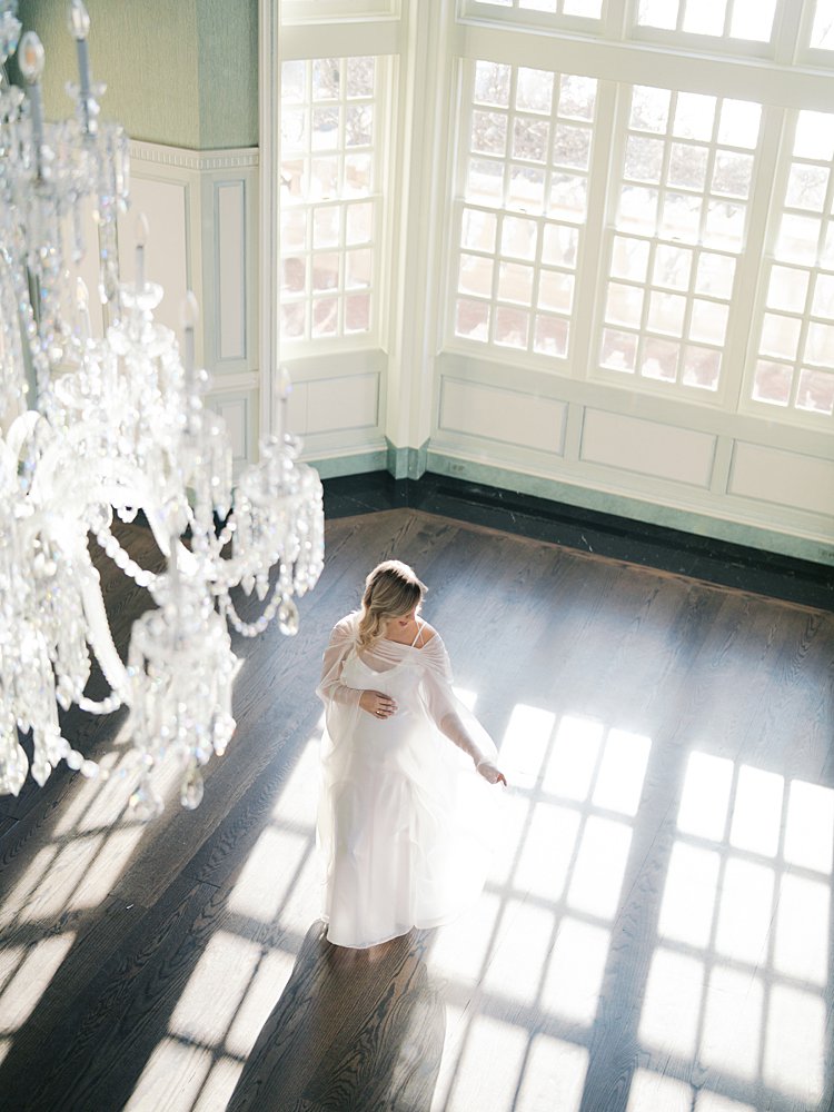 Pregnant Woman In White Dress Stands In Light In Front Of Large Windows And Chandelier At The Estate At River Run In Virginia.