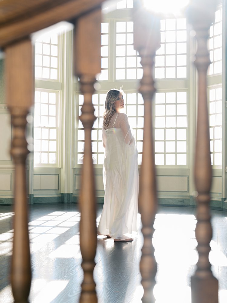 Blonde Pregnant Woman Stands In Front Of Large Windows During Her Maternity Photos At The Estate At River Run.