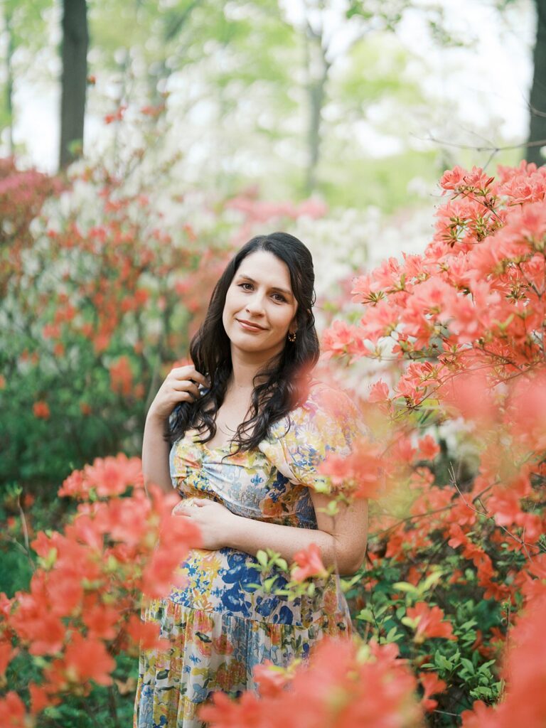 A Glowing Pregnant Woman In A Colorful Floral Dress Smiles Softly At The Camera, Surrounded By Lush Coral And Red Azaleas In Full Bloom During Her Clarksville Maternity Photos Session At Brighton Dam Azalea Garden