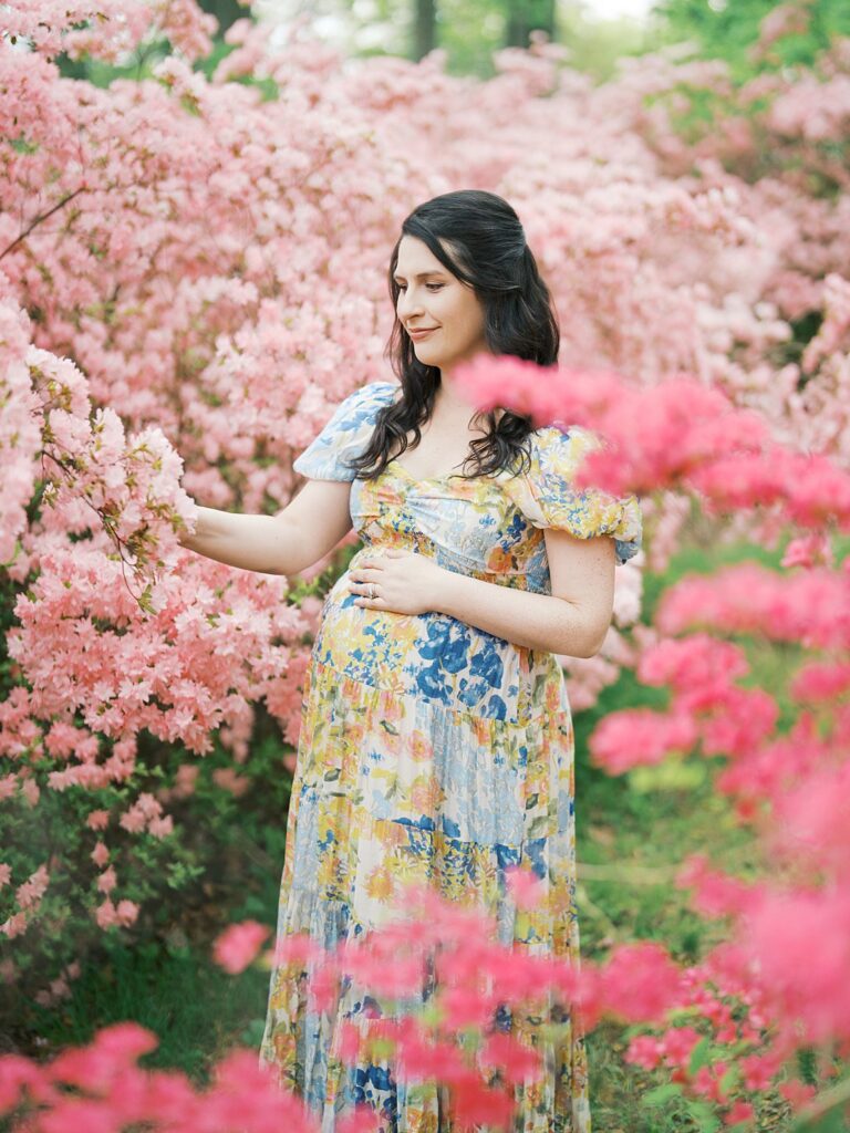 A Glowing Pregnant Woman In A Floral Maxi Dress Gently Touches A Branch Of Pink Azalea Blooms, Pausing To Look Down At Her Baby Bump In A Serene And Ethereal Clarksville Maternity Photos Moment At Brighton Dam Azalea Garden