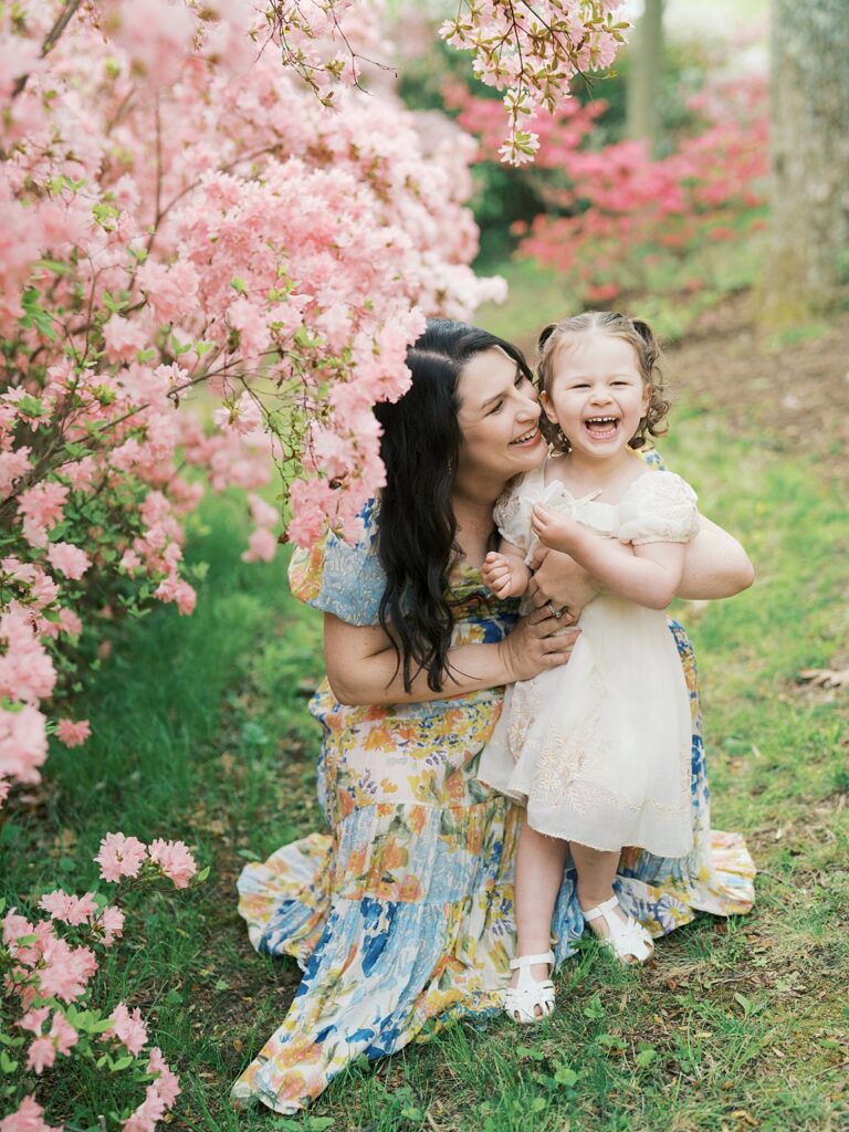A Pregnant Mother In A Colorful Floral Dress Crouches Beneath A Canopy Of Pink Azalea Blooms, Laughing With Her Delighted Toddler Daughter At Brighton Dam Azalea Garden During Their Clarksville Maternity Photos Session.