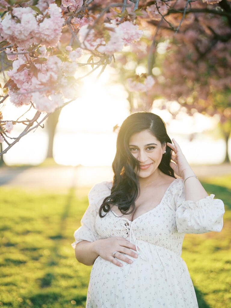 Pregnant Mother Smiling Softly Beneath Blooming Pink Cherry Blossoms At Hains Point, With Golden Sunlight Glowing Behind Her.