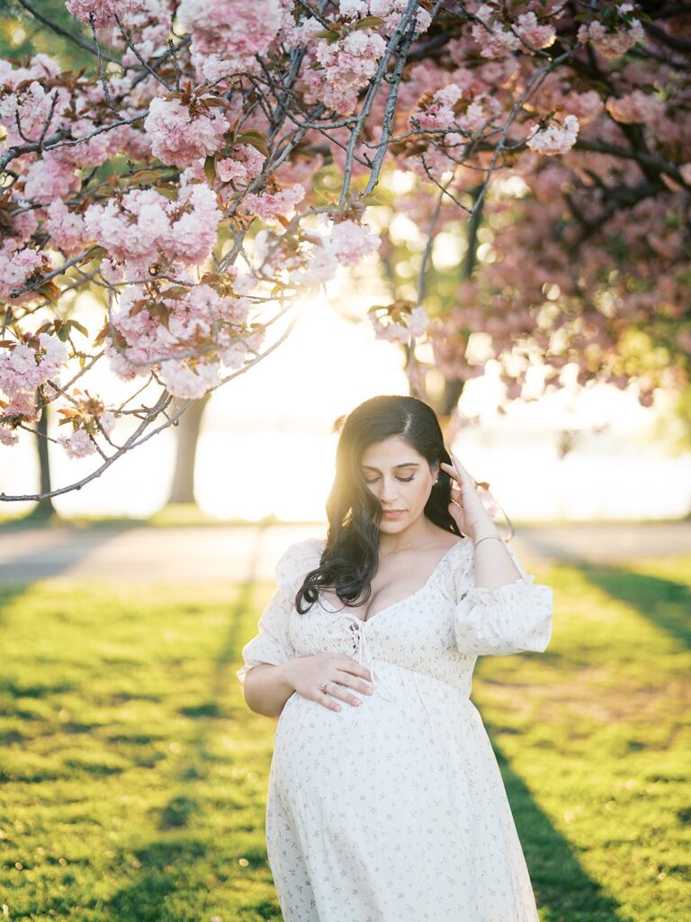 Pregnant Mother Standing Beneath Pink Cherry Blossoms At Hains Point, Softly Touching Her Hair As Golden Sunlight Filters Through The Trees Behind Her.