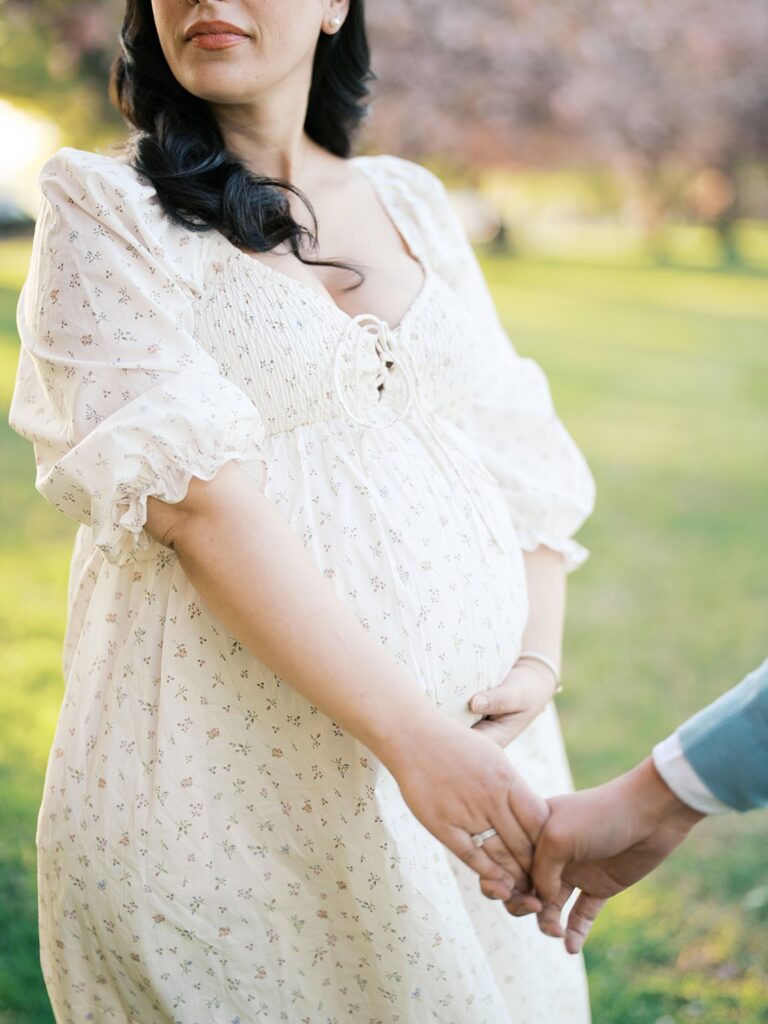 Close-Up Of An Expectant Mother Holding Hands With Her Partner, Her Baby Bump Gently Cradled In Soft Spring Light At East Potomac Park.