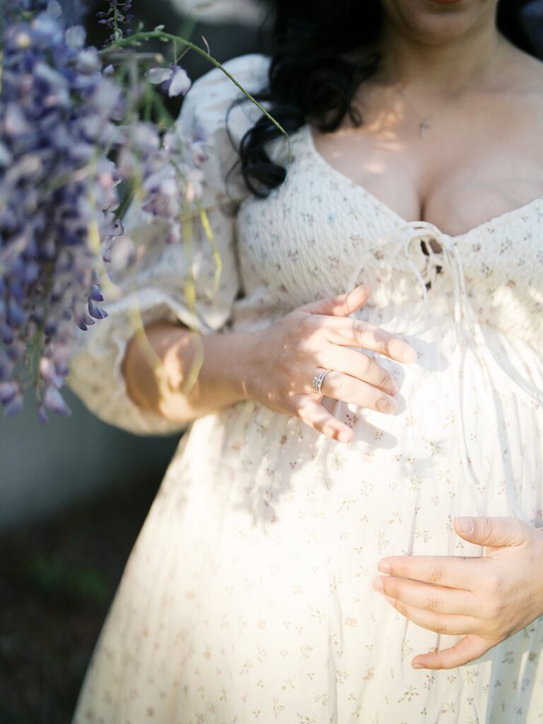 Intimate Detail Of A Pregnant Mother’s Hands Resting On Her Bump, Framed By Soft Purple Blossoms During Dc Wisteria Maternity Photos.