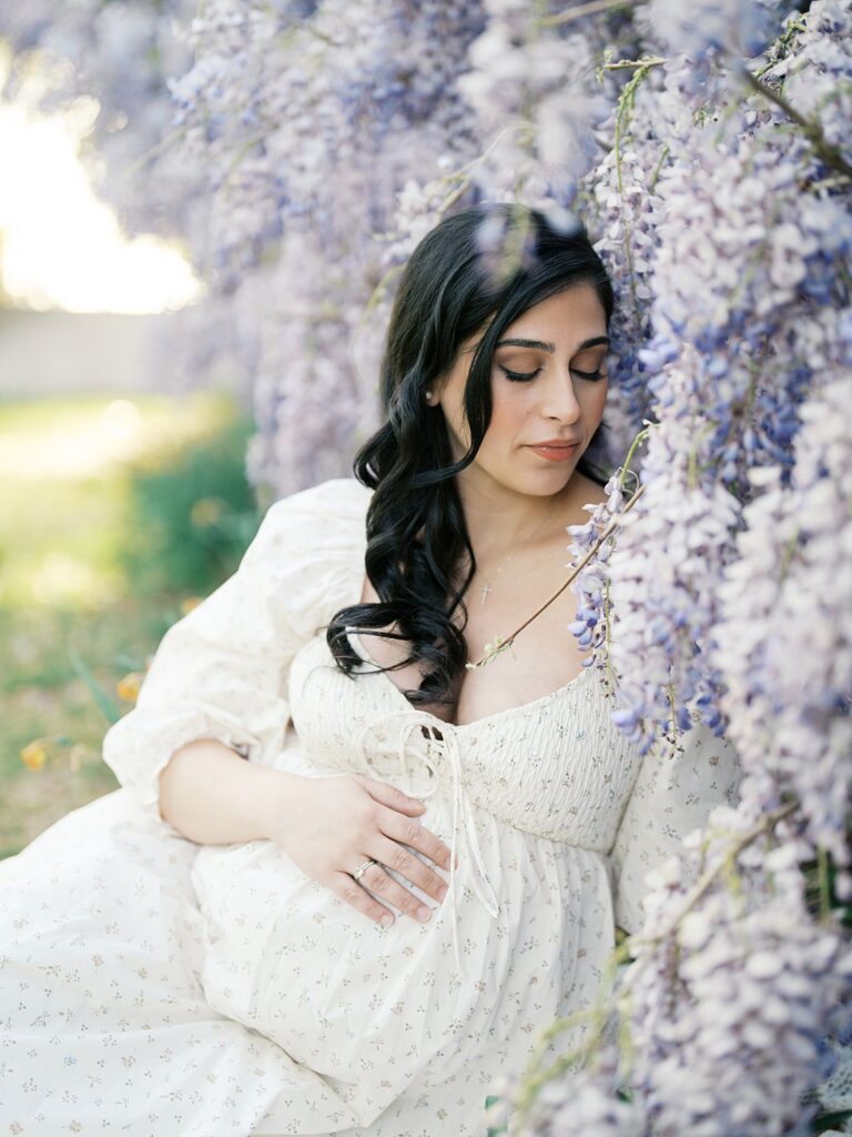 Maternity Portrait Of A Mother Seated Under Cascading Lavender Wisteria, Softly Cradling Her Bump During Dc Wisteria Maternity Photos With Warm Golden Light.