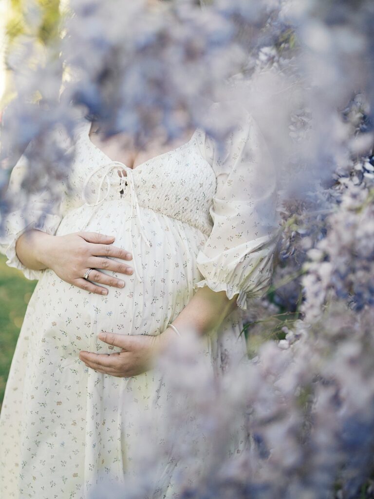 Close-Up Of A Pregnant Mother’s Hands Resting On Her Belly, Framed By Blurred Wisteria Blossoms During Dc Wisteria Maternity Photos.