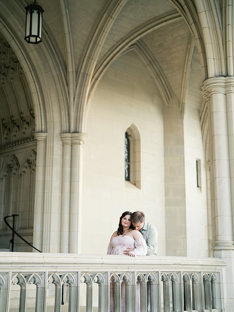 Couple Embracing Under Vaulted Arches During Maternity Photos At The National Cathedral Cherry Blossoms, Blending Soft Emotion With Timeless Architectural Details.