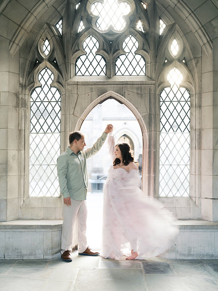 Couple Dancing Beneath Arched Stone Windows Inside The Cathedral Grounds During Maternity Photos At The National Cathedral Cherry Blossoms, Blending Architecture With Movement And Connection.