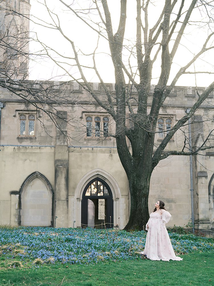 Expectant Mother Standing In A Field Of Blue Flowers With Cathedral Architecture Behind Her During Maternity Photos At The National Cathedral Cherry Blossoms, Creating A Peaceful, Airy Scene.