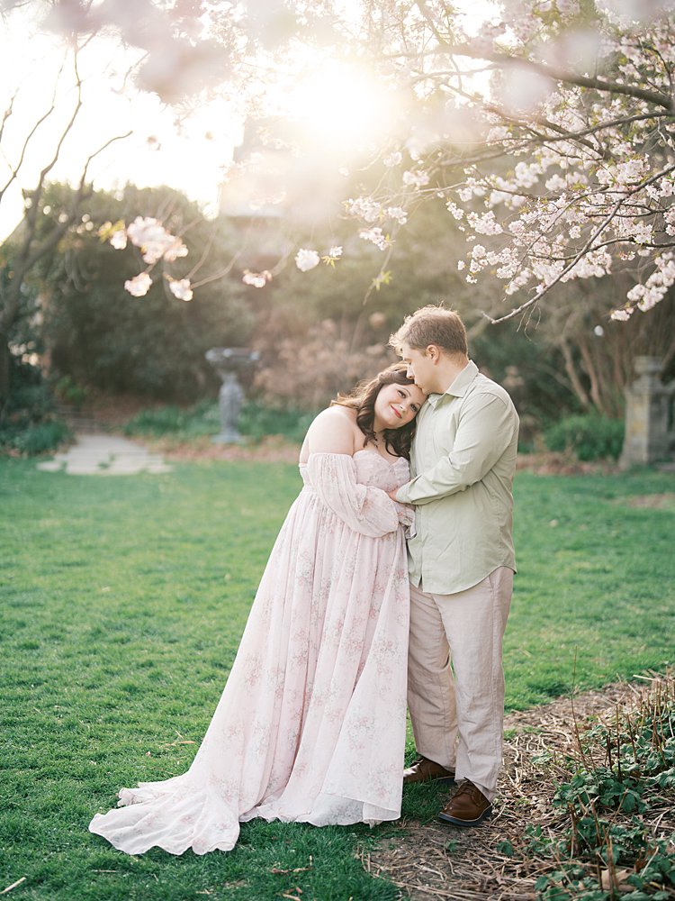 Couple Embracing Under Glowing Evening Light And Blooming Branches During Maternity Photos At The National Cathedral Cherry Blossoms, Capturing A Quiet, Intimate Moment.