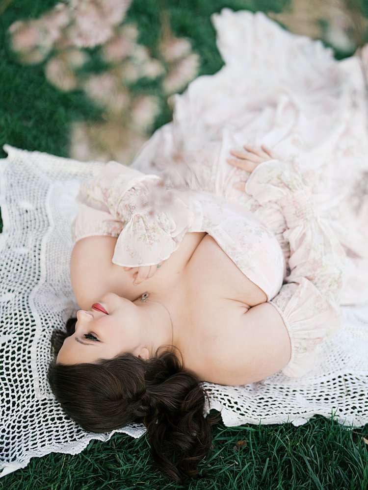 Pregnant Mother Lying On A Knit Blanket In A Flowing Dress During Maternity Photos At The National Cathedral Cherry Blossoms, Captured In A Soft And Romantic Overhead Portrait.