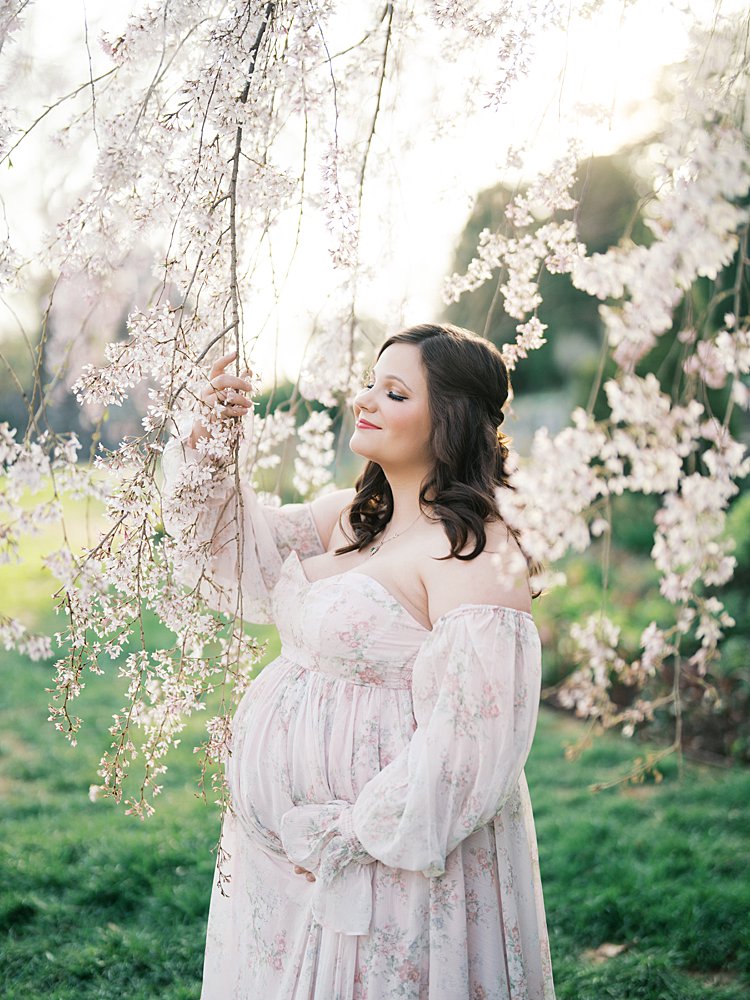 Expectant Mother Gently Holding Cherry Blossom Branches During Maternity Photos At The National Cathedral Cherry Blossoms, Surrounded By Soft Pink Blooms And Glowing Light.