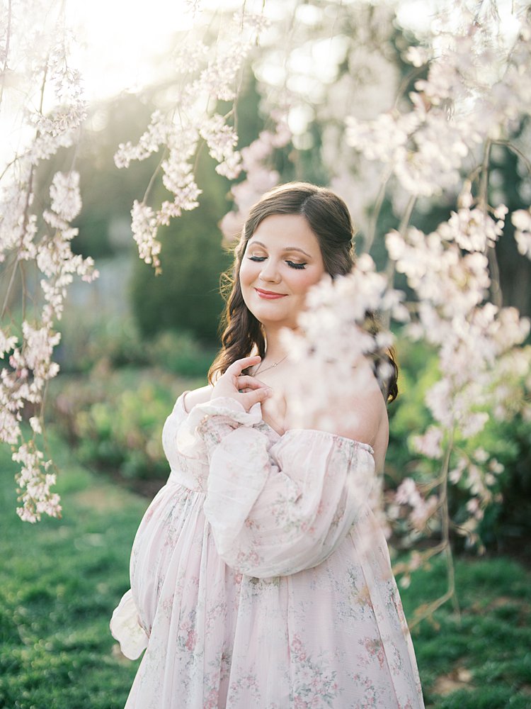 Expectant Mother In A Flowing Blush Gown Standing Among Soft Pink Blooms During Maternity Photos At The National Cathedral Cherry Blossoms, With Dreamy Light And Delicate Florals Framing Her Face.