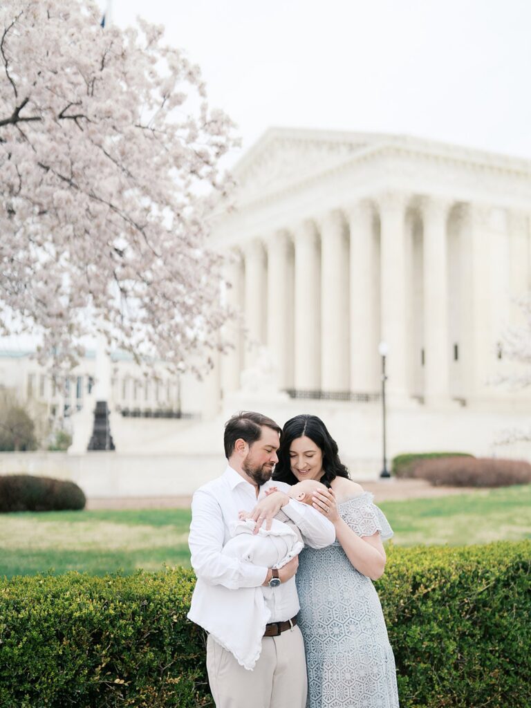 A Mother And Father Huddle Close Over Their Newborn Baby On The Lawn In Front Of The United States Supreme Court Building, Framed By Blooming Cherry Blossom Trees During A Washington Dc Newborn Photography Session.