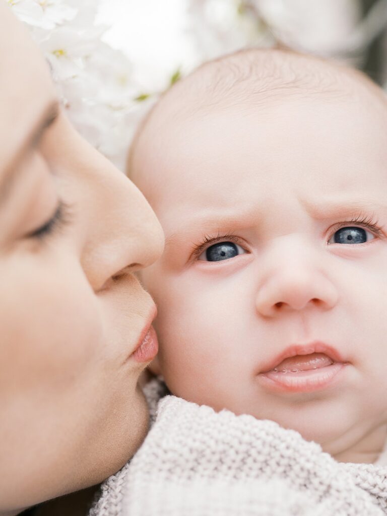 A Close-Up Of A Mother Gently Kissing Her Wide-Eyed Newborn Baby On The Cheek, The Baby Wrapped In A Soft Cream Knit Blanket With Delicate Cherry Blossoms Softly Blurred In The Background.