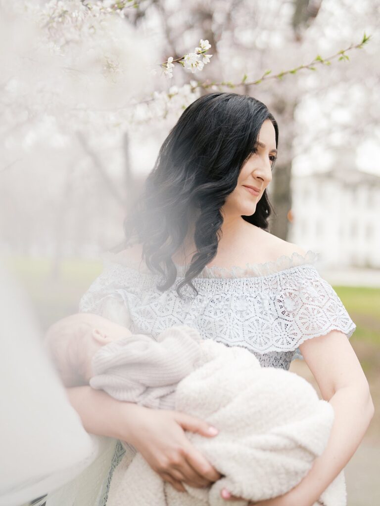 A Dark-Haired Mother In A White Lace Off-The-Shoulder Dress Smiles Softly While Cradling Her Sleeping Newborn Baby, With Cherry Blossom Trees Softly Blurred In The Background During A Stanton Park Newborn Session