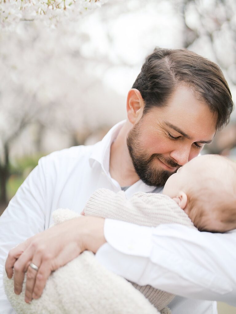 A Bearded Father In A White Button-Down Shirt Leans Down To Nuzzle His Sleeping Newborn Baby, His Eyes Closed In A Tender Moment Captured During A Stanton Park Cherry Blossoms Newborn Session