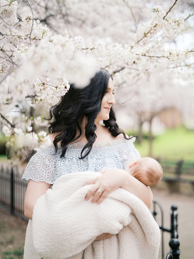A Mother With Long Dark Curly Hair Stands In Profile Beneath Blooming Cherry Blossom Trees At Stanton Park, Holding Her Newborn Baby Close During Their Spring Newborn Photo Session In Dc