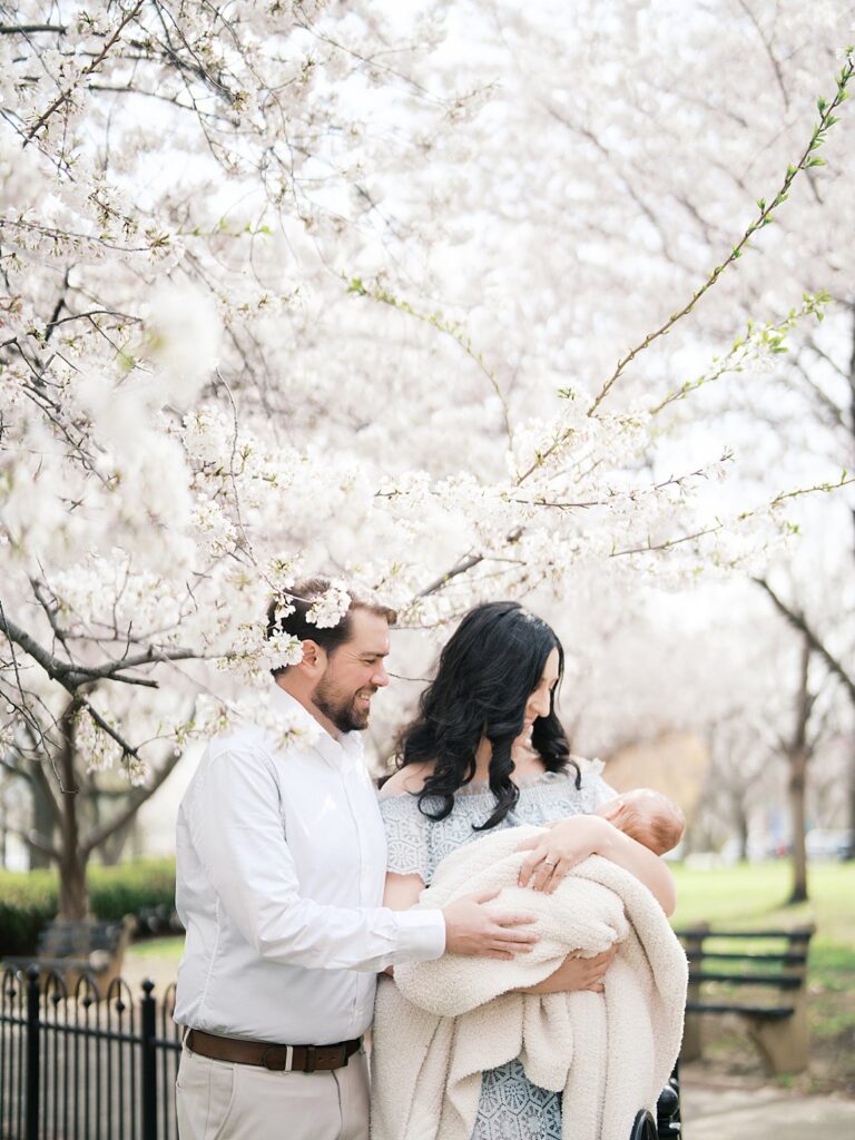 A Mother And Father Lean In Close Over Their Bundled Newborn Baby Beneath The Stanton Park Cherry Blossoms, The Mother Wearing A Floral Crown And Soft Blue Dress During Their Newborn Photos.