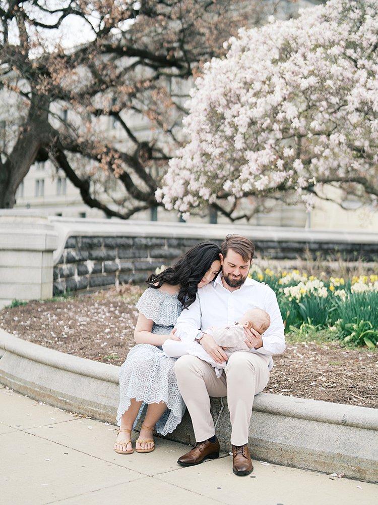 A Couple Sits Together On A Stone Ledge Surrounded By Blooming Cherry Blossom Trees, Gazing Down At Their Newborn Wrapped In A Cream Blanket — A Beautiful Example Of What Happens After Your Session When Fleeting Moments Become Timeless Portraits.