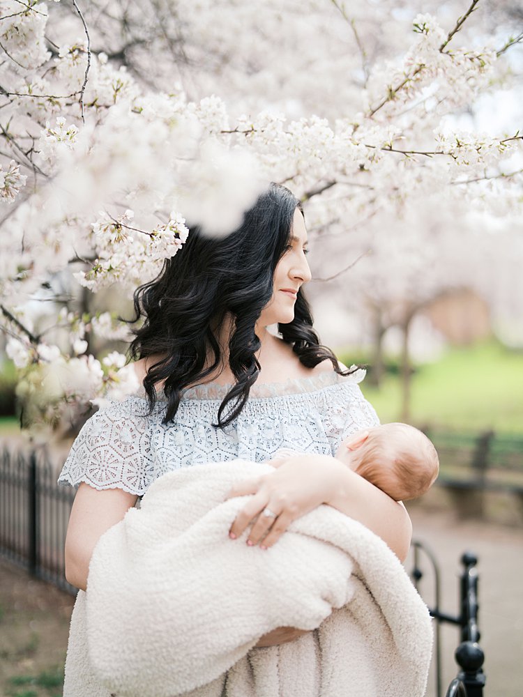 A Dark-Haired Mother In A Blue Lace Dress Holds Her Newborn Swaddled In A Cream Knit Blanket, Smiling Softly Beneath A Canopy Of White Cherry Blossoms During An Outdoor Spring Newborn Session.