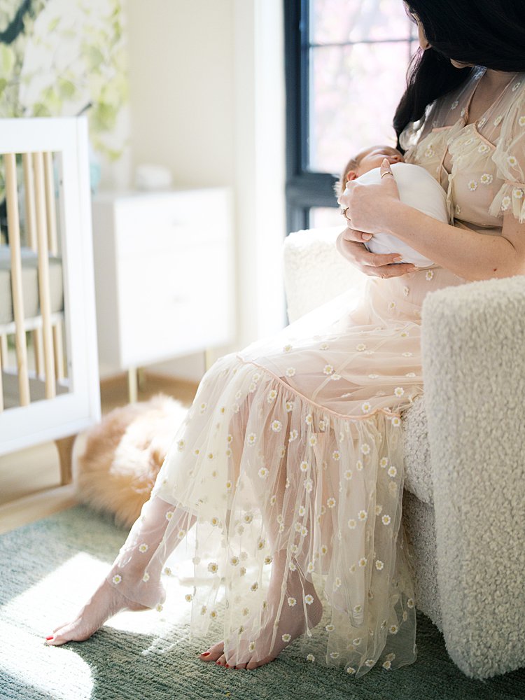 A Close-Up Of A Mother In A Flowing Blush Floral Dress Seated In A Sunlit Nursery, Holding Her Newborn In Her Lap — A Tender In-Home Moment That Captures What Happens After Your Session When The Images Finally Arrive.