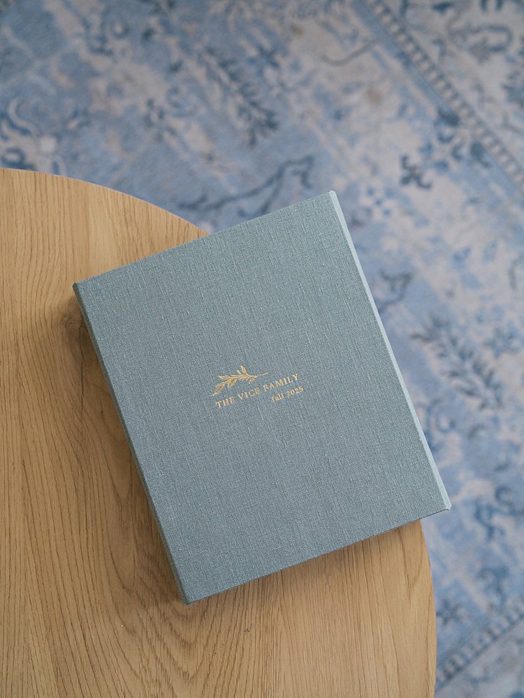 A Dusty Blue Linen Heirloom Album Personalized With Gold Foil Family Name And Date Sits On A Warm Wood Surface — A Timeless Keepsake That Shows What Happens After Your Session And How Your Portraits Are Preserved For Generations.