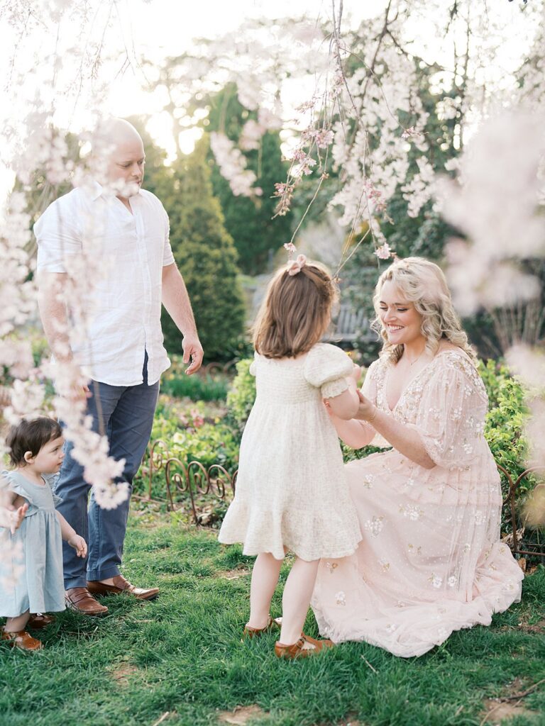A Family Gathers Beneath Delicate Spring Blossoms As Parents And Young Children Share A Quiet Moment Together, A Beautiful Example Of When Is The Best Time To Get Family Photos In The Dmv With Soft Florals In Bloom.