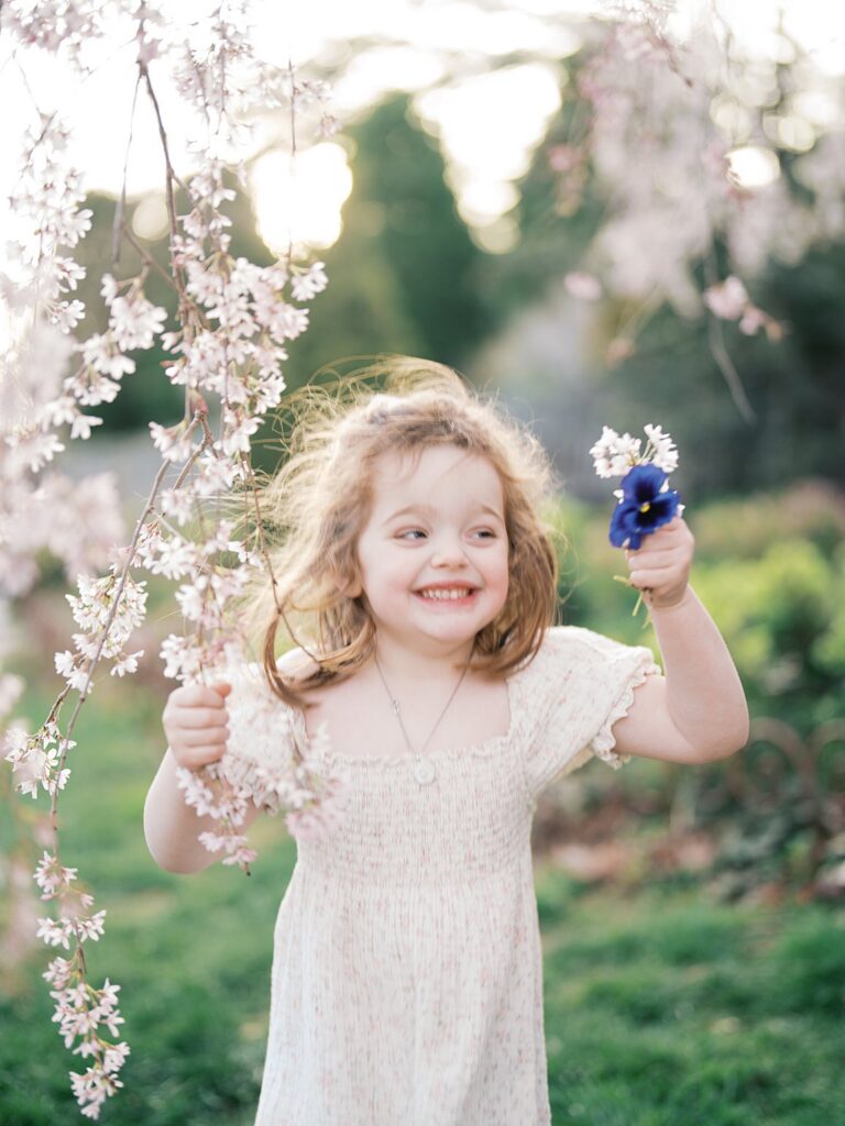 A Joyful Little Girl Smiles While Holding A Flower Beneath Hanging Spring Blooms, Surrounded By Soft Greenery And Pastel Tones That Feel Like The Perfect Answer To When Is The Best Time To Get Family Photos In The Dmv.