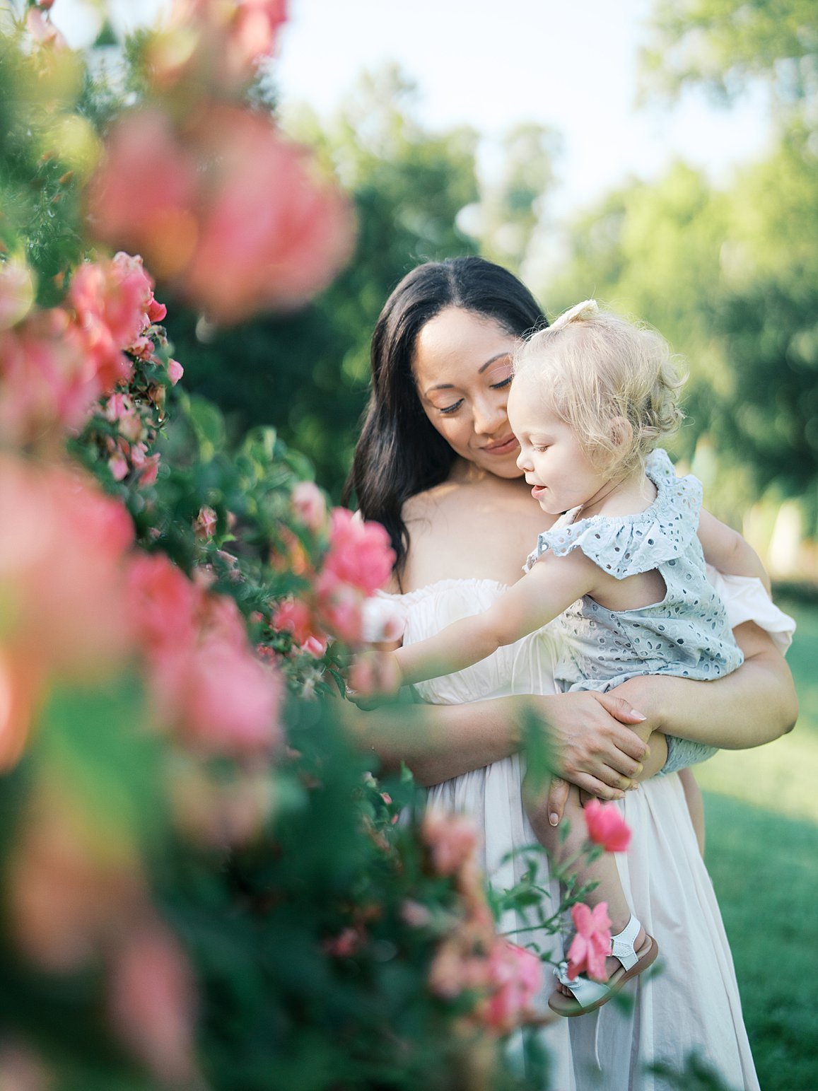 A Mother Holds Her Child While They Gently Explore Blooming Garden Roses, Surrounded By Lush Greenery And Soft Light, A Beautiful Reflection Of When Is The Best Time To Get Family Photos In The Dmv.