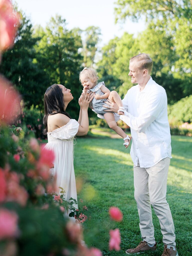 A Joyful Family Moment As Parents Lift Their Toddler Into The Air In A Sunlit Garden, The Vibrant Greens And Florals Highlighting When Is The Best Time To Get Family Photos In The Dmv For Bright, Playful Images.
